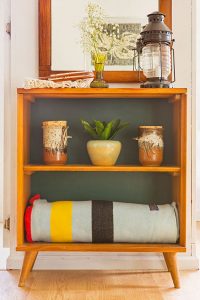 A small Mid Century Modern bookshelf with a blanket, pottery and a lantern in it.
