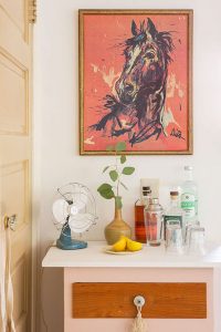 A vintage pink cabinet has been turned into a bar station. A horse art print hangs above on the wall.