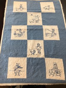 This blanket has two colors: muted blue and off-white. It has six off-white squares, each depicting a child or fairytale character.