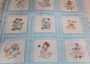 This vintage quilt has white squares embroidered with clothed kittens, framed by pale blue fabric.