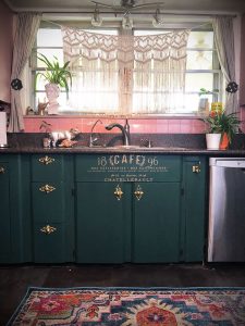 A vintage-inspired kitchen set up with dark green cabinets with "cafe" written in gold lettering upon the center one.
