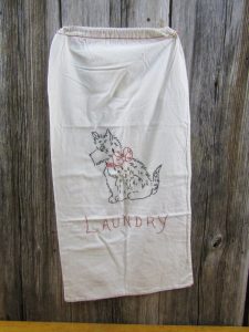 This white laundry bag has a stitched black Scottie dog with a red bow above the word "laundry" in red.
