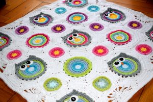 This crocheted baby blanket is white with colorful modern owl motifs that alternate with colorful circles.