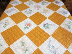 This quilt has mustard-yellow and white squares with state flowers stitched behind their embroidered state names.