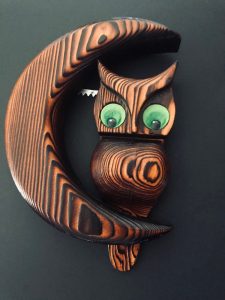 This wooden wall art is an owl with large green eyes perched on the crook of a crescent moon.