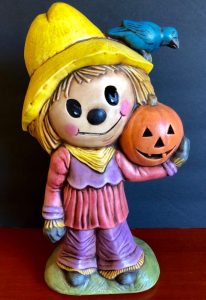 A small painted statue of a girl scarecrow dressed in pink and purple with a yellow rain-hat holding a jack-o-lantern.