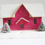 Glittery, magenta putz house with snow covered roof and two mini decorated trees in front of it.