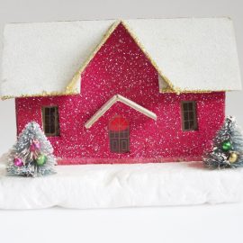 Glittery, magenta putz house with snow covered roof and two mini decorated trees in front of it.