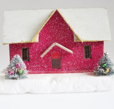 Glittery, magenta putz house with snow covered roof and two mini decorated trees in front of it.