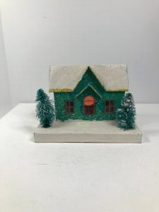 Green putz house with a snowy roof and two mini trees in front of it.
