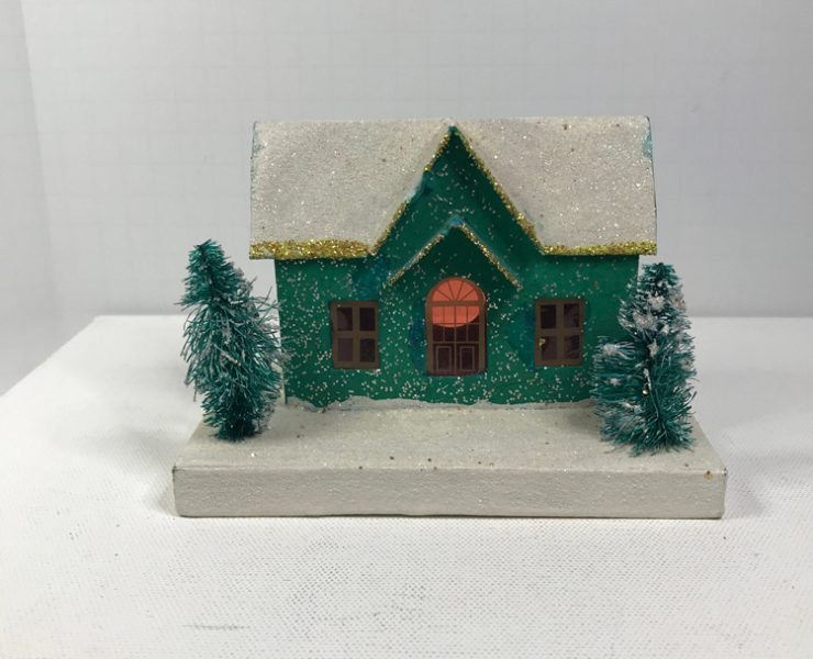 Green putz house with a snowy roof and two mini trees in front of it.