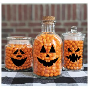 Different shaped glass jars with black jack-o-lantern face decals and orange jelly beans inside.