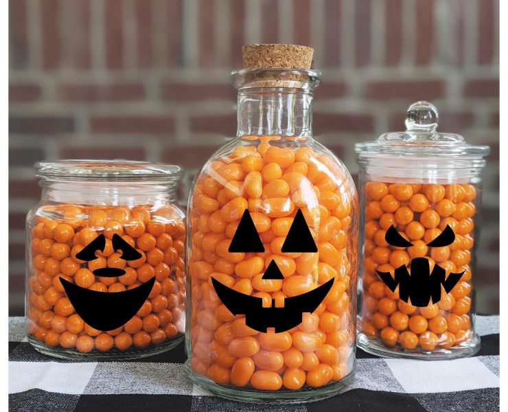 Different shaped glass jars with black jack-o-lantern face decals and orange jelly beans inside.