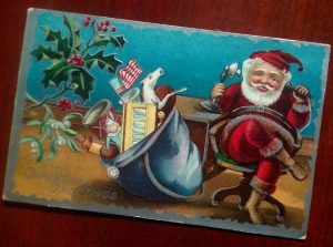 Postcard with Santa sitting at a desk talking on the telephone with his bag of toys beside him on the floor.