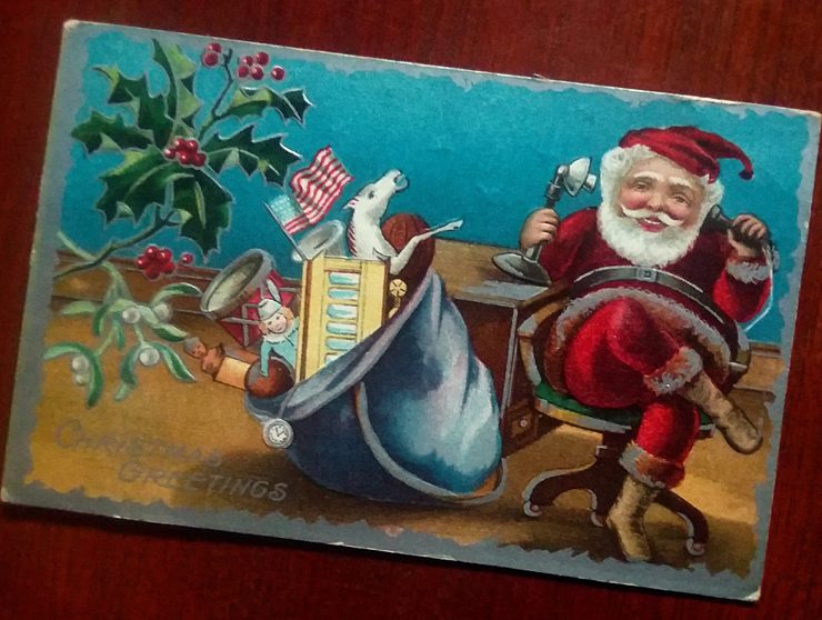 Postcard with Santa sitting at a desk talking on the telephone with his bag of toys beside him on the floor.