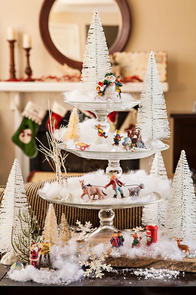 Three cake stands are stacked to create a winter scene with snow, bottlebrush trees and miniature figurines.