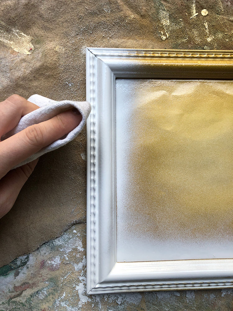 a frame is spray painted in a layer of gold and white. Part of the top layer is being removed by cloth.